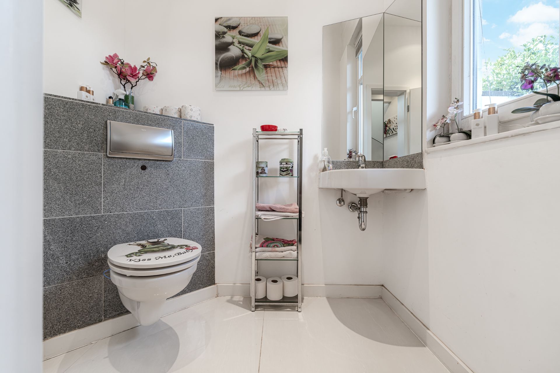 Modernes Badezimmer mit Wandtoilette, Waschbecken, Regal und Fenster mit Blick nach draußen.
