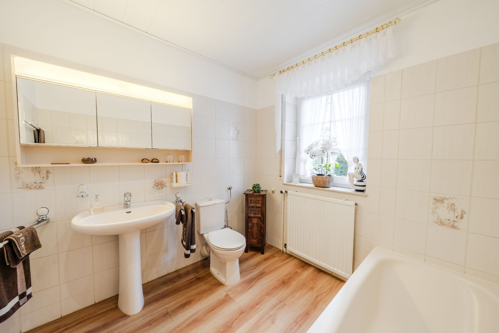 Helles Badezimmer mit Waschbecken, Toilette, Badewanne und Fenster mit Vorhang.