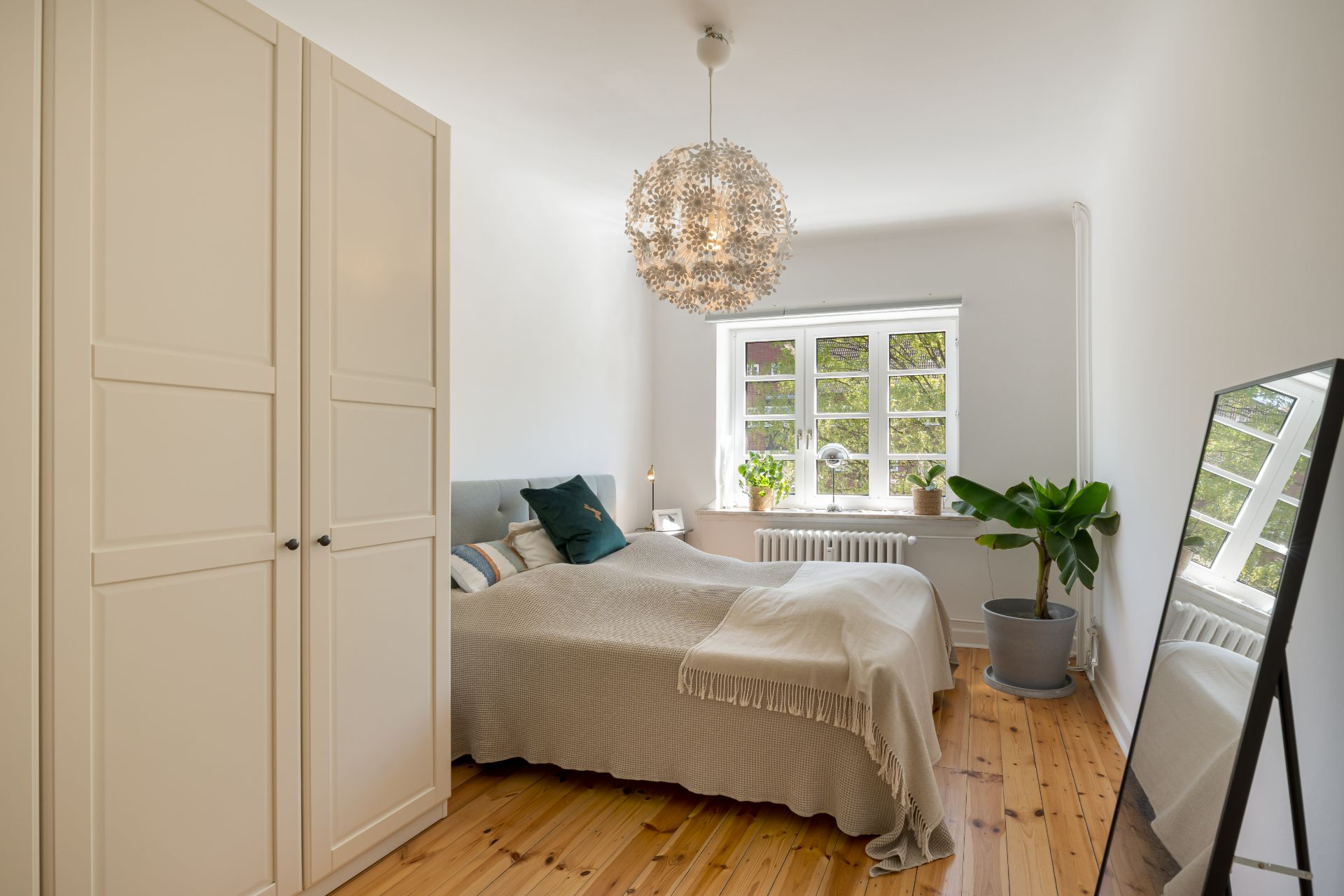 Helles Schlafzimmer mit Doppelbett, Kleiderschrank, großem Fenster, Stehspiegel und Zimmerpflanze.
