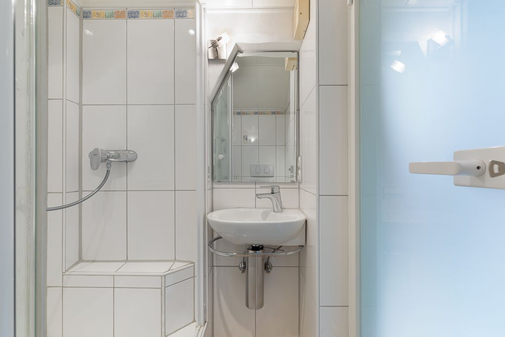 Kleines Badezimmer mit Dusche, Waschbecken und Spiegel, weiße Fliesen an den Wänden.