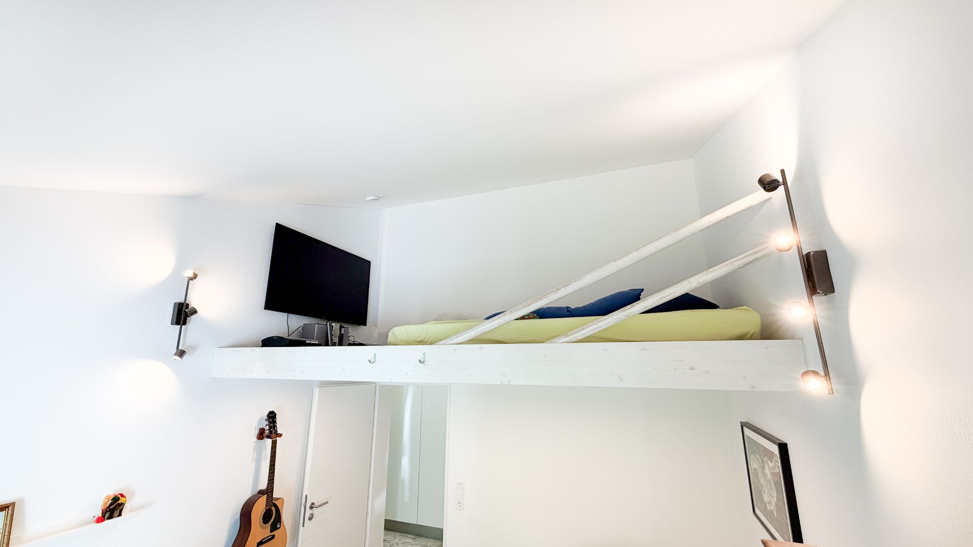 Helles Zimmer mit Hochbett, Wandlampen, Gitarre und Fernseher an weißer Wand.