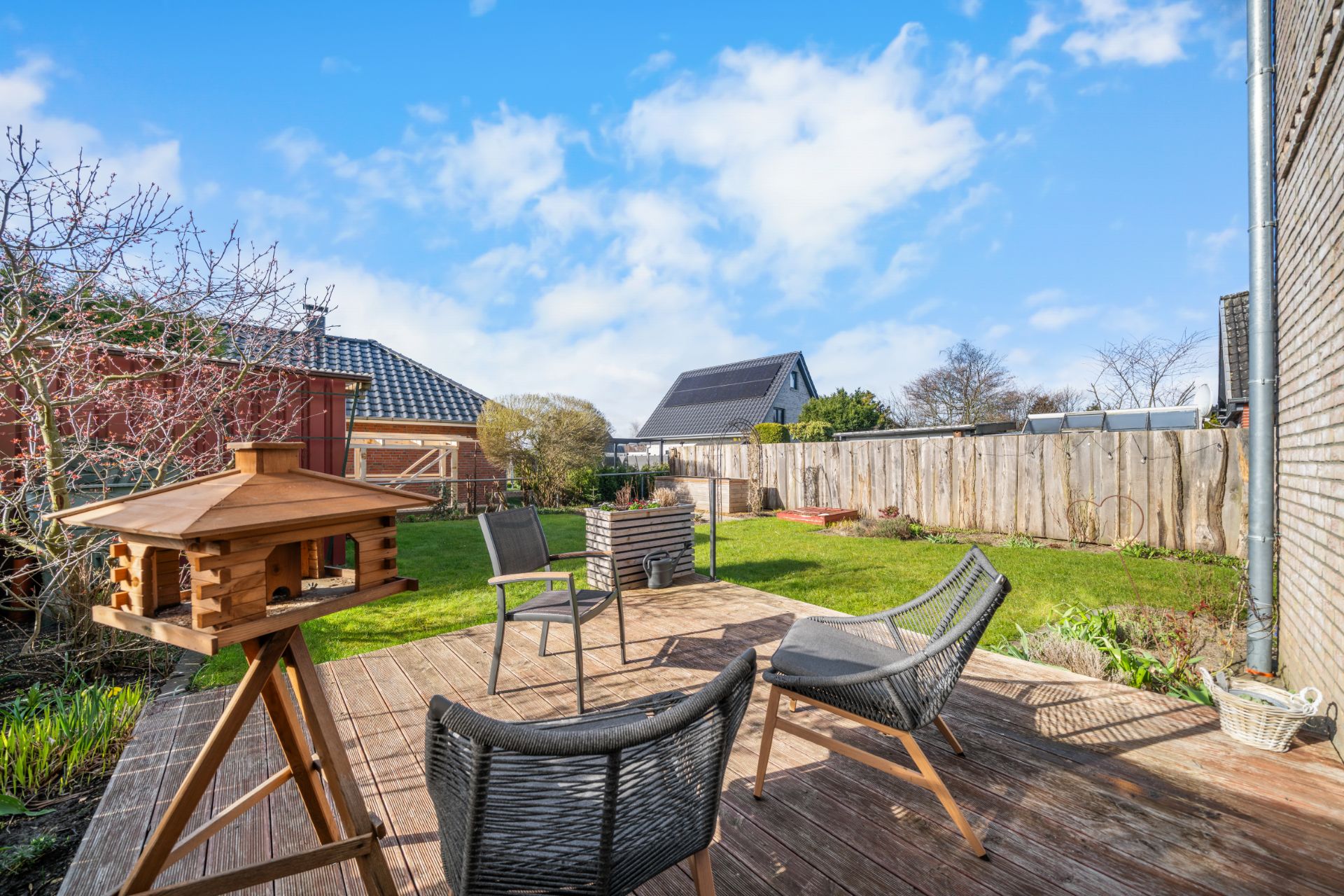 Holzterrasse mit Stühlen, Vogelhaus und Blick auf Garten mit Zaun.