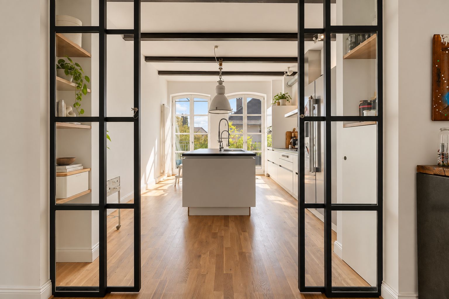 Moderne helle Küche mit Kücheninsel, Holzboden und großen Fenstern, durch schwarze Glastüren sichtbar.