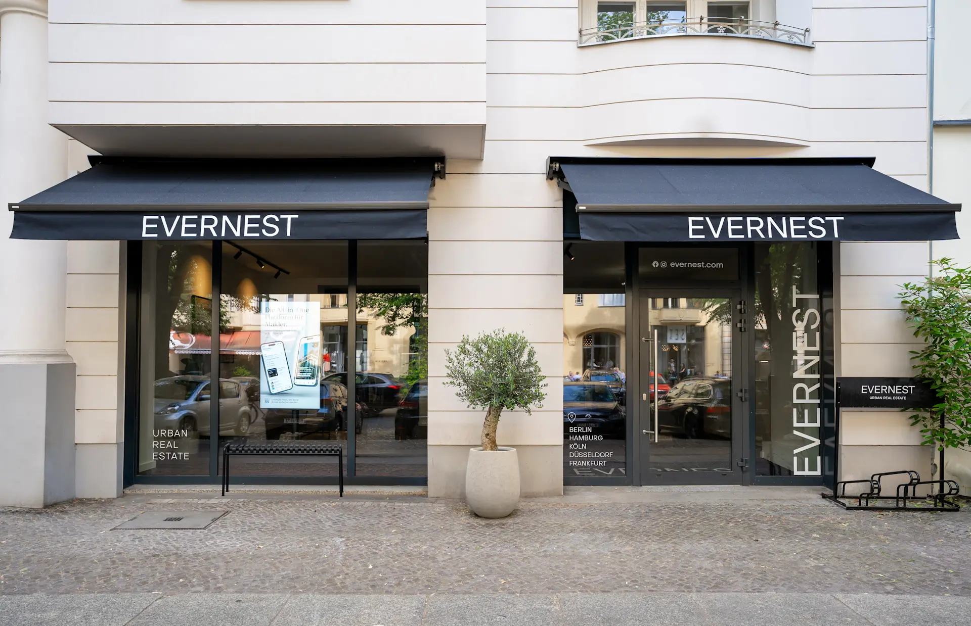Geschäft mit schwarzen Markisen und großer Glasfront, ein Olivenbaum im Topf steht draußen.