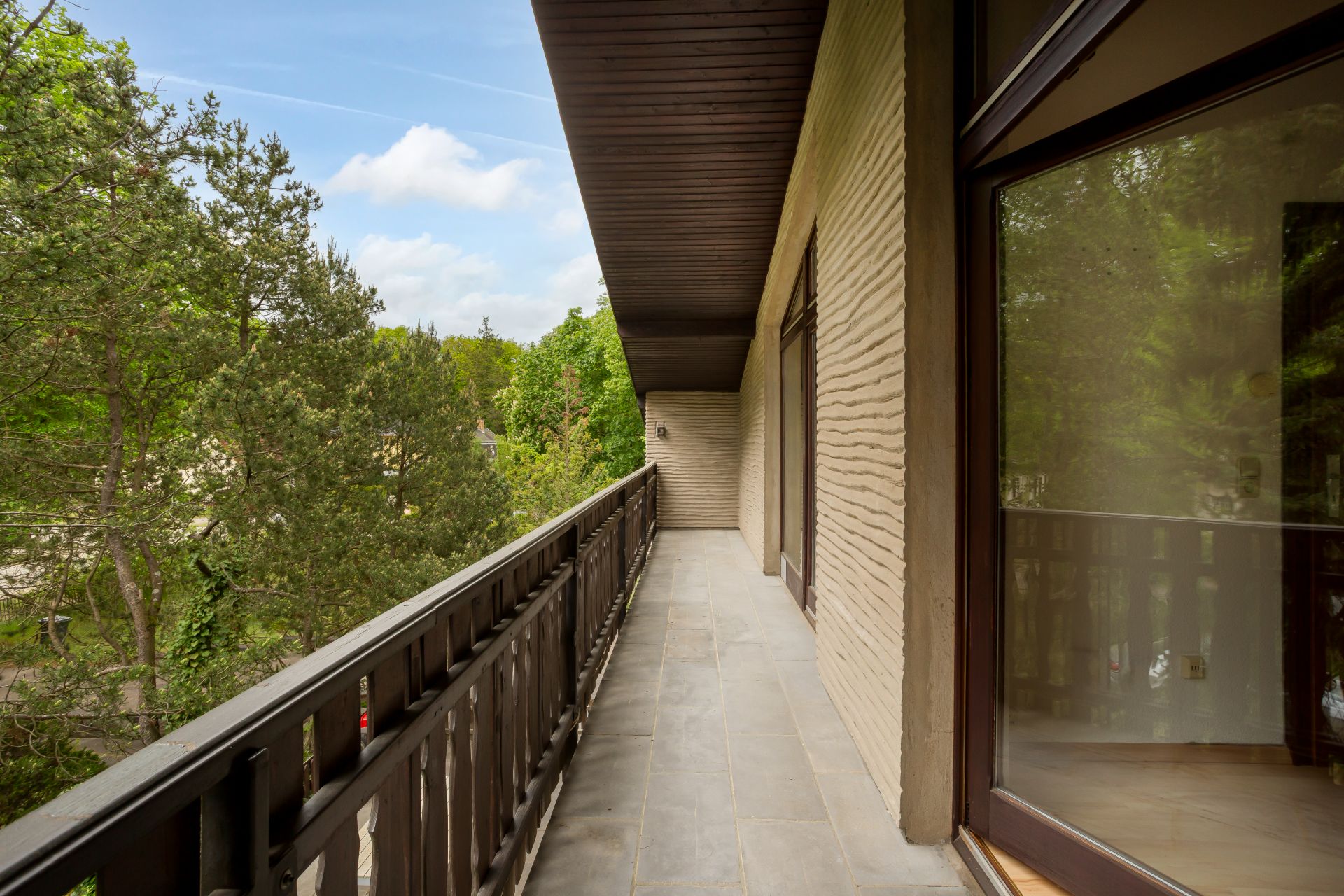 Langer Balkon mit Holzgeländer und Blick auf grüne Bäume.