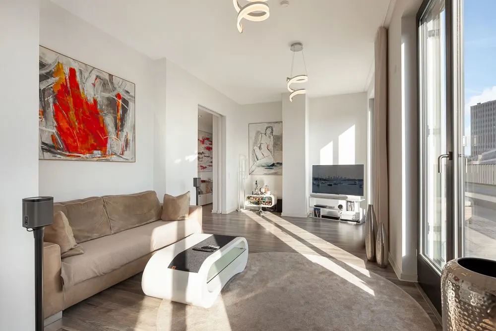 Modernes Wohnzimmer mit beiger Couch, Couchtisch, großem Fenster und Fernseher auf Holzfußboden.