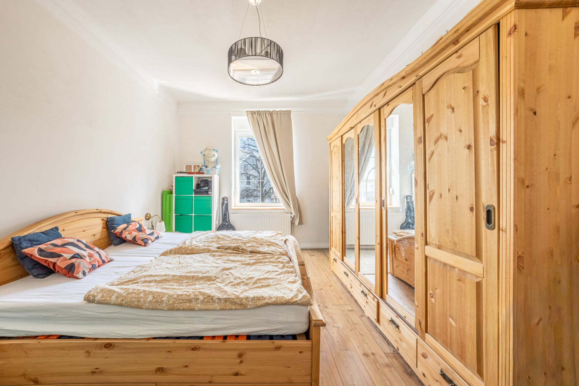 Helles Schlafzimmer mit Holzbett, großem Kleiderschrank und Fenster mit beiger Gardine.