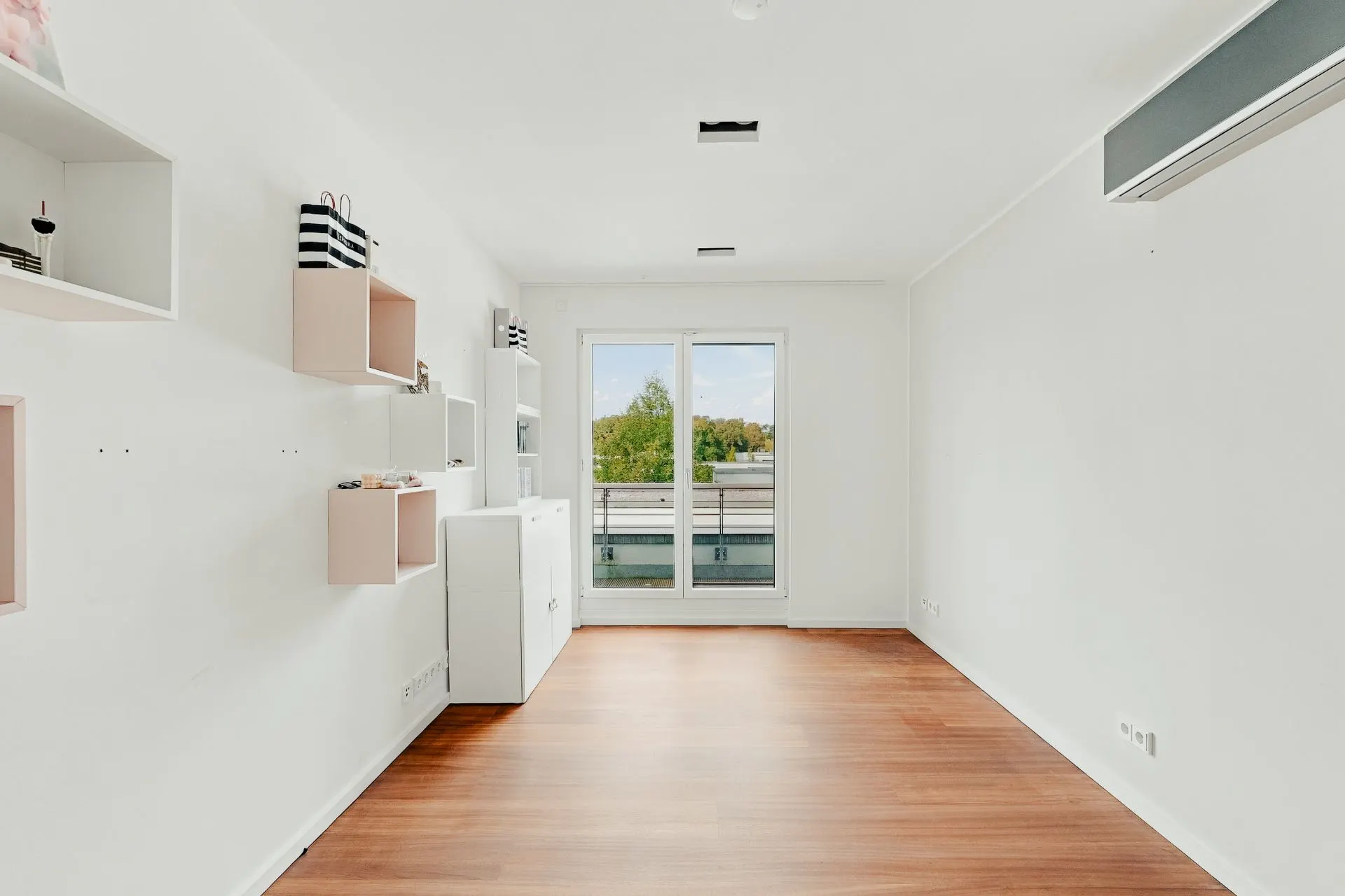 Heller Raum mit Holzfußboden, weißen Wänden, Regalen und Fenster mit Blick ins Grüne.