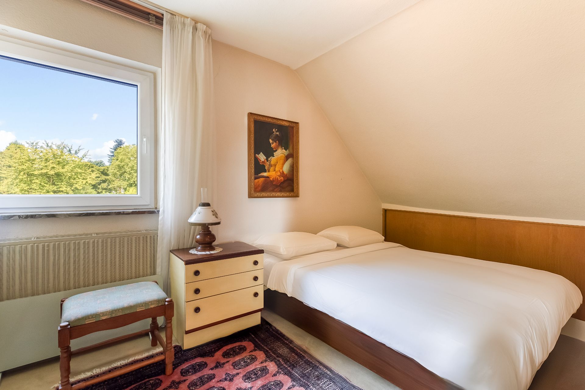 Kleines Schlafzimmer mit Doppelbett, Nachttisch, Lampe, Stuhl und Fenster mit Blick ins Grüne.
