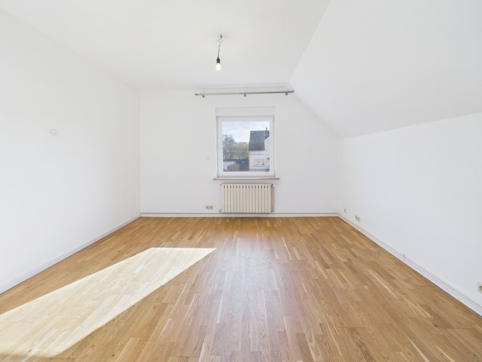 Helles, leeres Zimmer mit Holzboden, weiß gestrichenen Wänden und einem Fenster mit Heizung darunter.