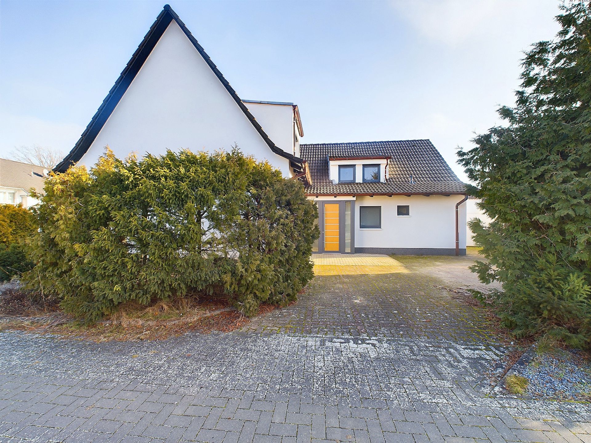 Einfamilienhaus mit dunklem Dach, gelber Haustür und Einfahrt, umgeben von Büschen.