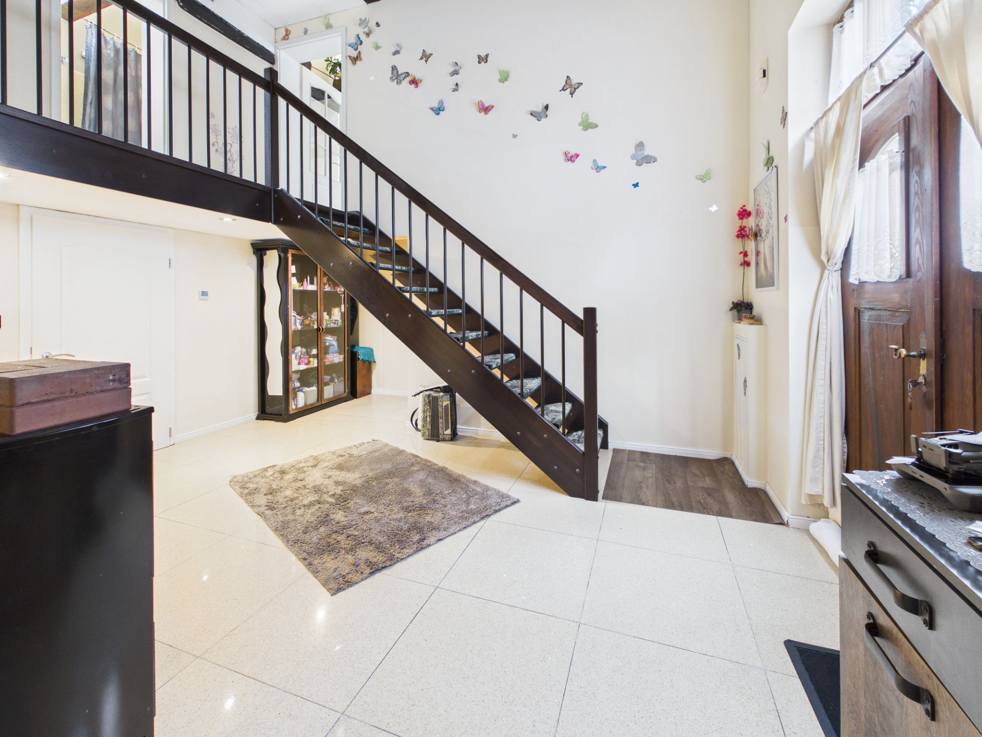Wohnzimmer mit offener Treppe, Teppich, Vitrine und Schmetterlings-Dekoration an der Wand.