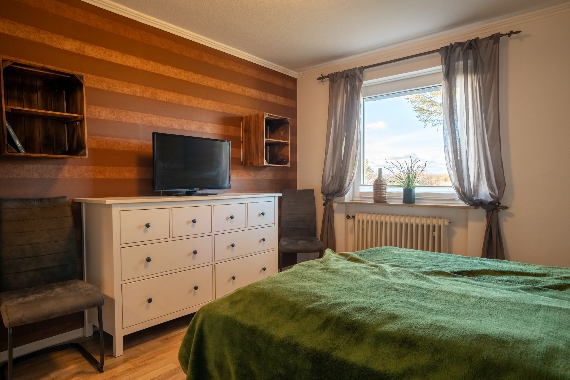 Schlafzimmer mit grünem Bett, Kommode, Fernseher, zwei Stühlen und Fenster mit Vorhängen.