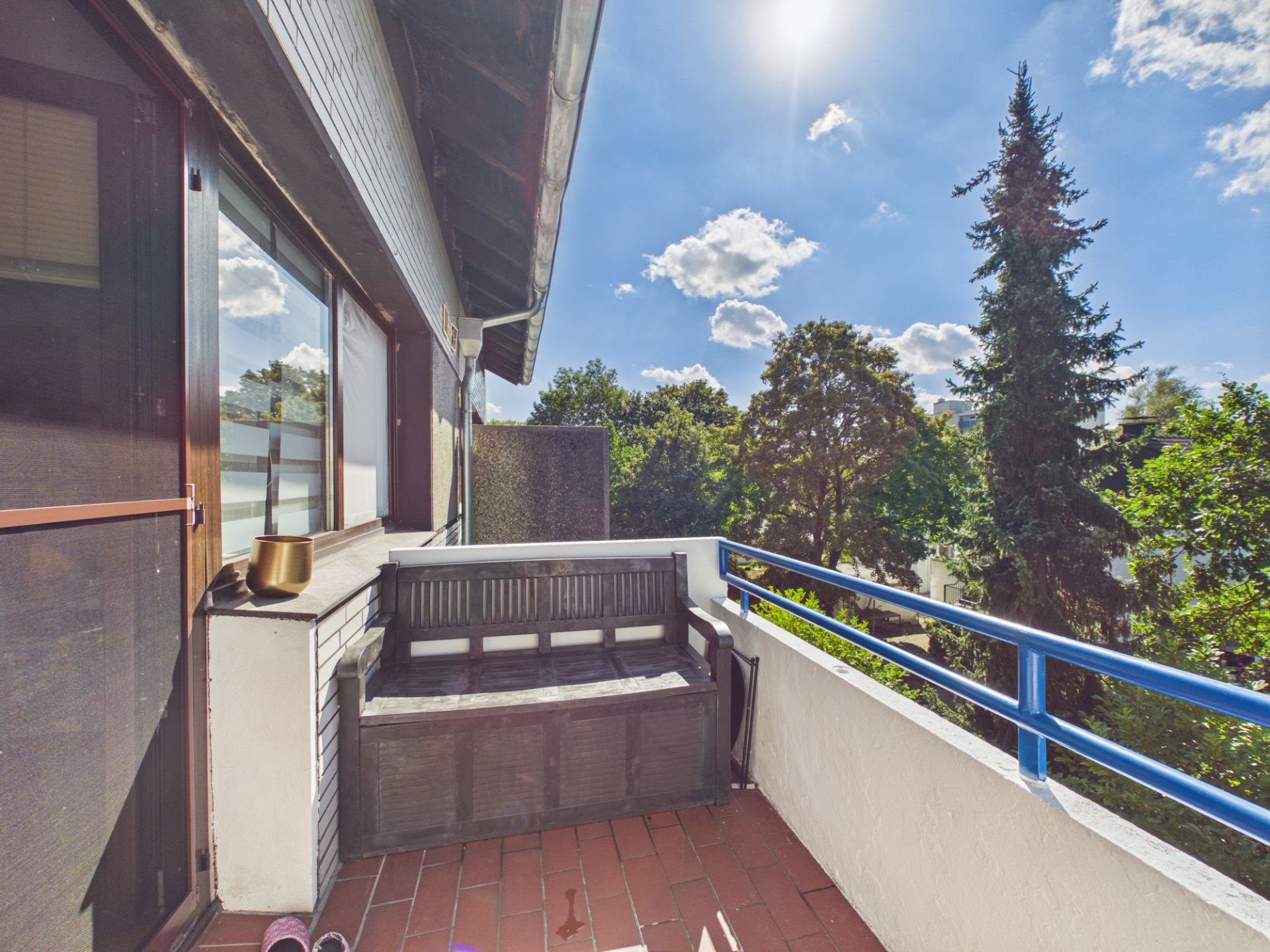 Balkon mit Bank, blauer Brüstung und Blick auf Bäume bei sonnigem Wetter.
