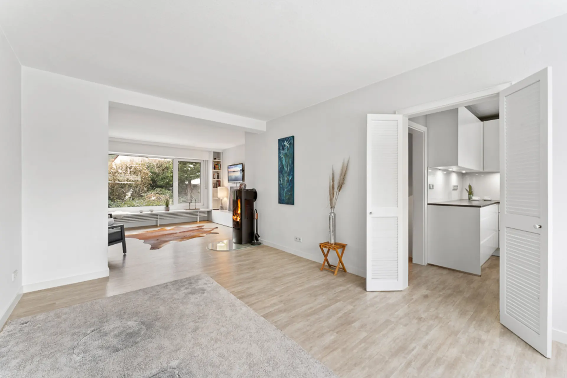 Helles Wohnzimmer mit Holzofen, großem Fenster, Teppich und angrenzender moderner Küche.