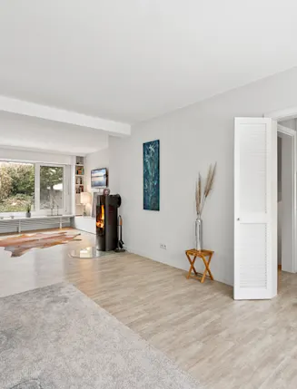 Helles Wohnzimmer mit Holzofen, großem Fenster, Teppich und angrenzender moderner Küche.