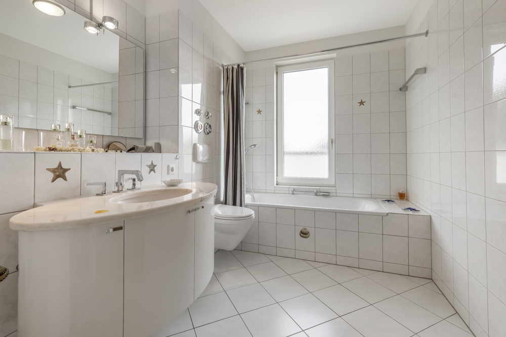 Helles Badezimmer mit Badewanne, großem Spiegel, Waschbecken und WC, alles in weiß gefliest.