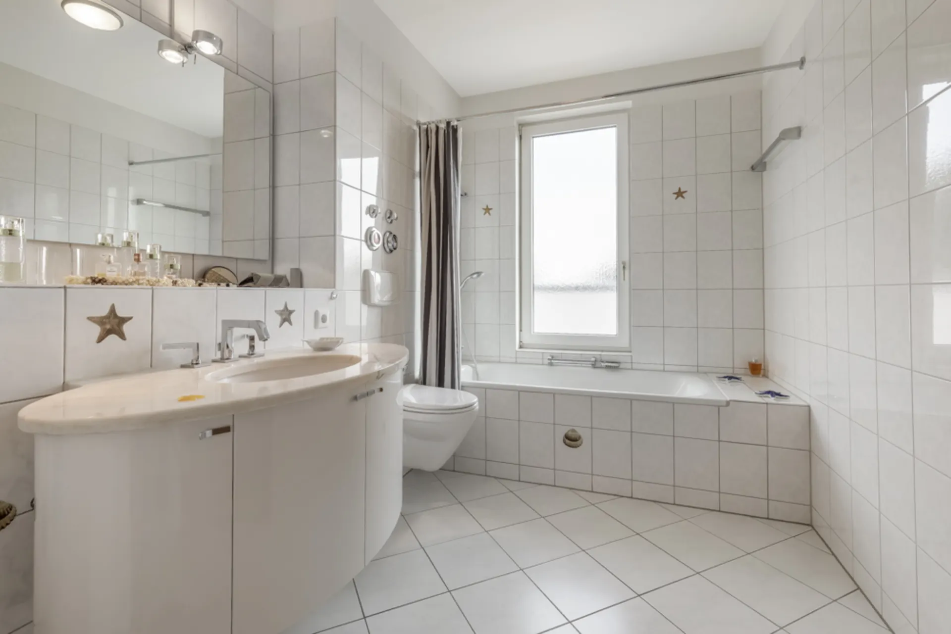Helles Badezimmer mit Badewanne, großem Spiegel, Waschbecken und WC, alles in weiß gefliest.