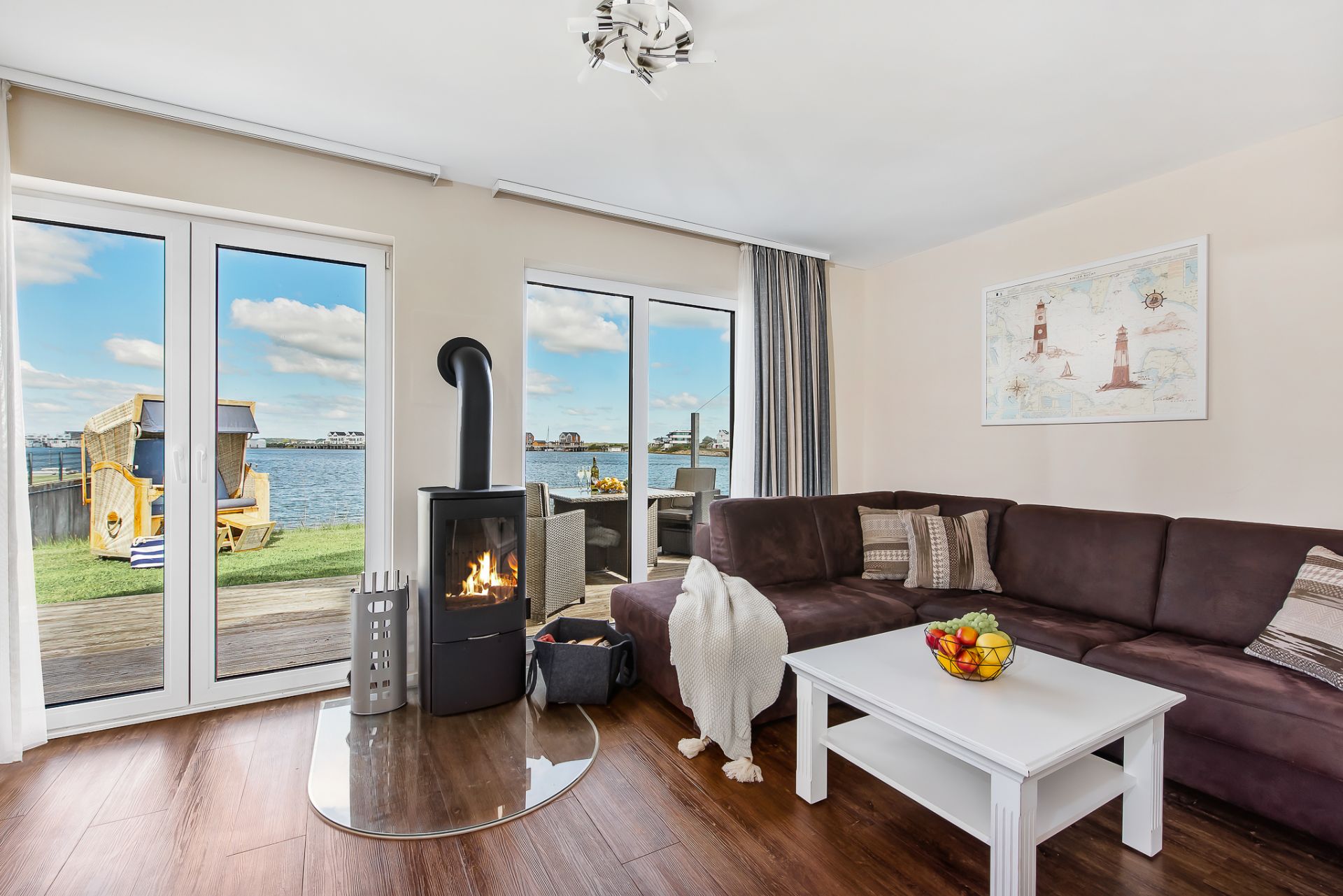 Wohnzimmer mit Ecksofa, Kamin, weißem Couchtisch und Blick auf Wasser durch große Fenster.
