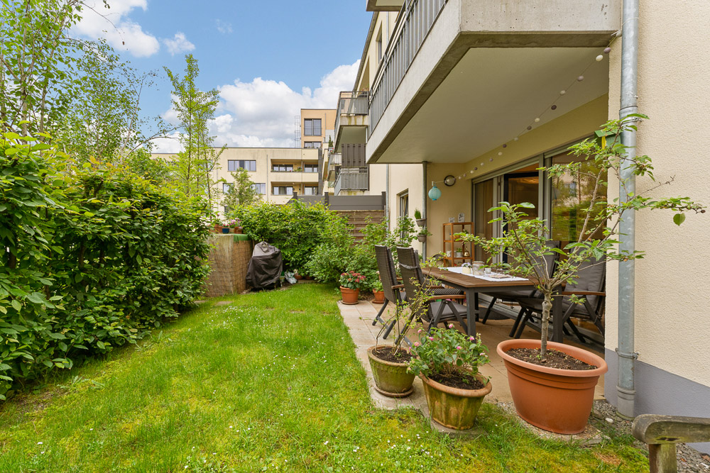 Terrasse mit Esstisch und Stühlen, umgeben von Pflanzen und Rasen im Garten eines Wohnhauses.