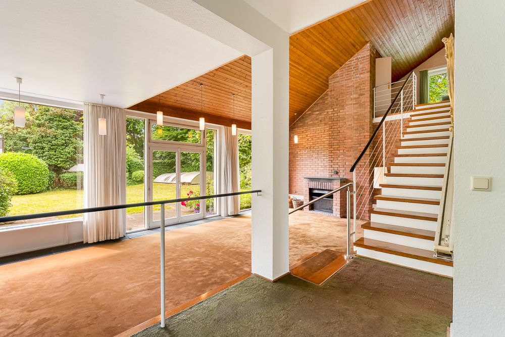 Wohnzimmer mit großen Fenstern, Kamin, Treppe und Blick in den Garten.