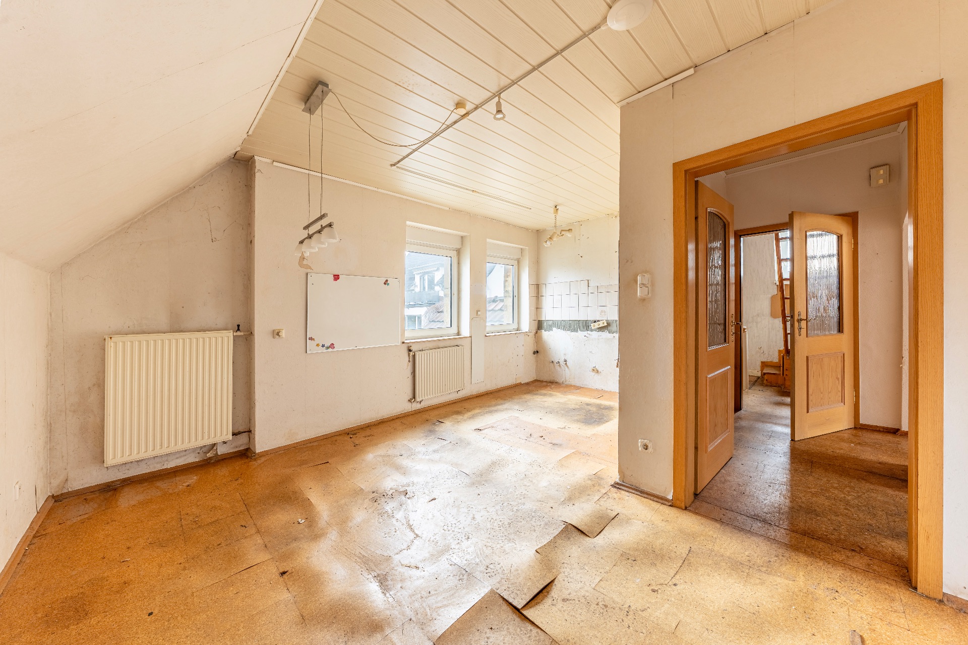 Leerer, heller Raum mit beschädigtem Boden, zwei Fenstern und Heizkörper in einem Wohnhaus.