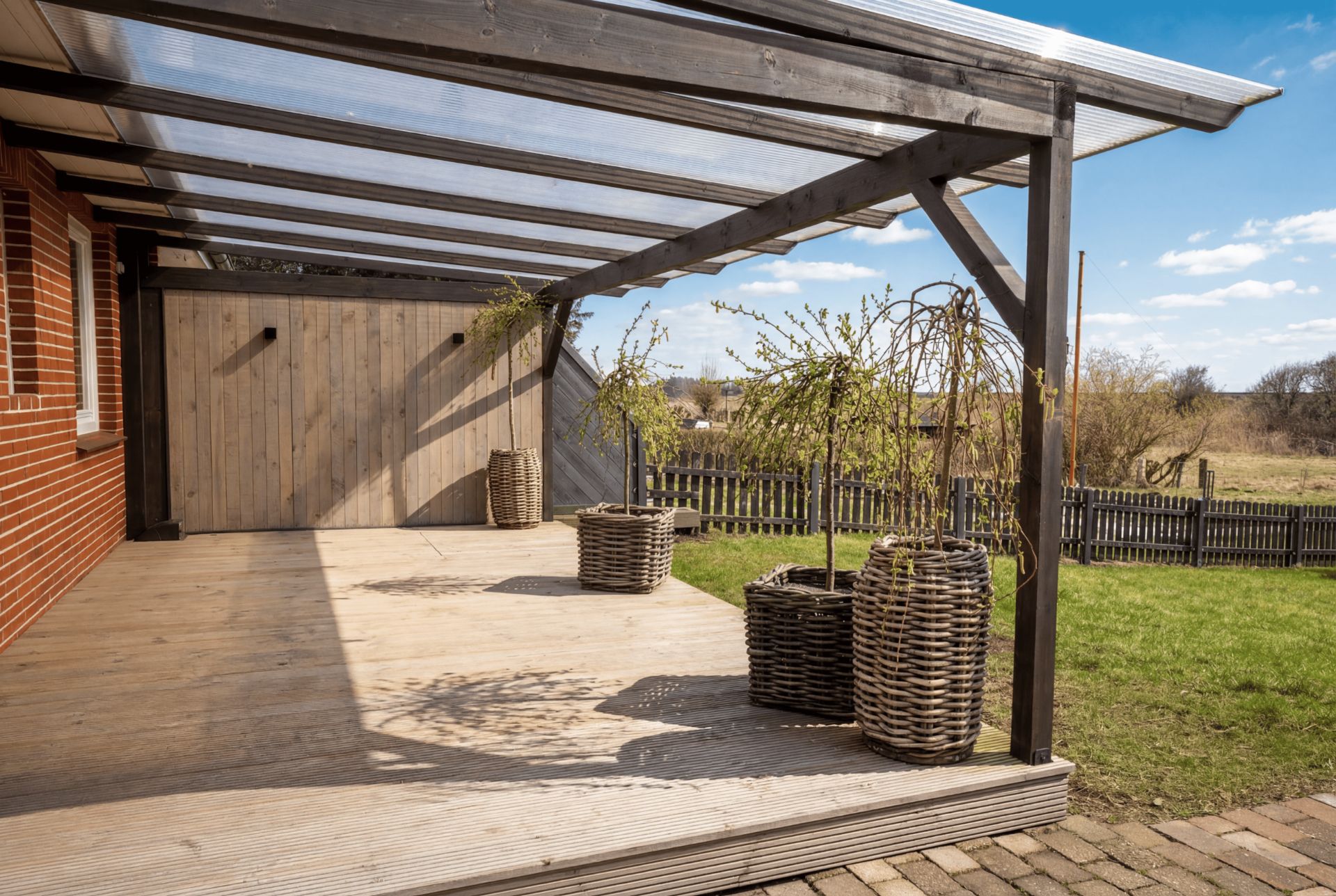 Überdachte Holzterrasse mit Korb-Blumentöpfen und Blick auf Garten und Zaun.