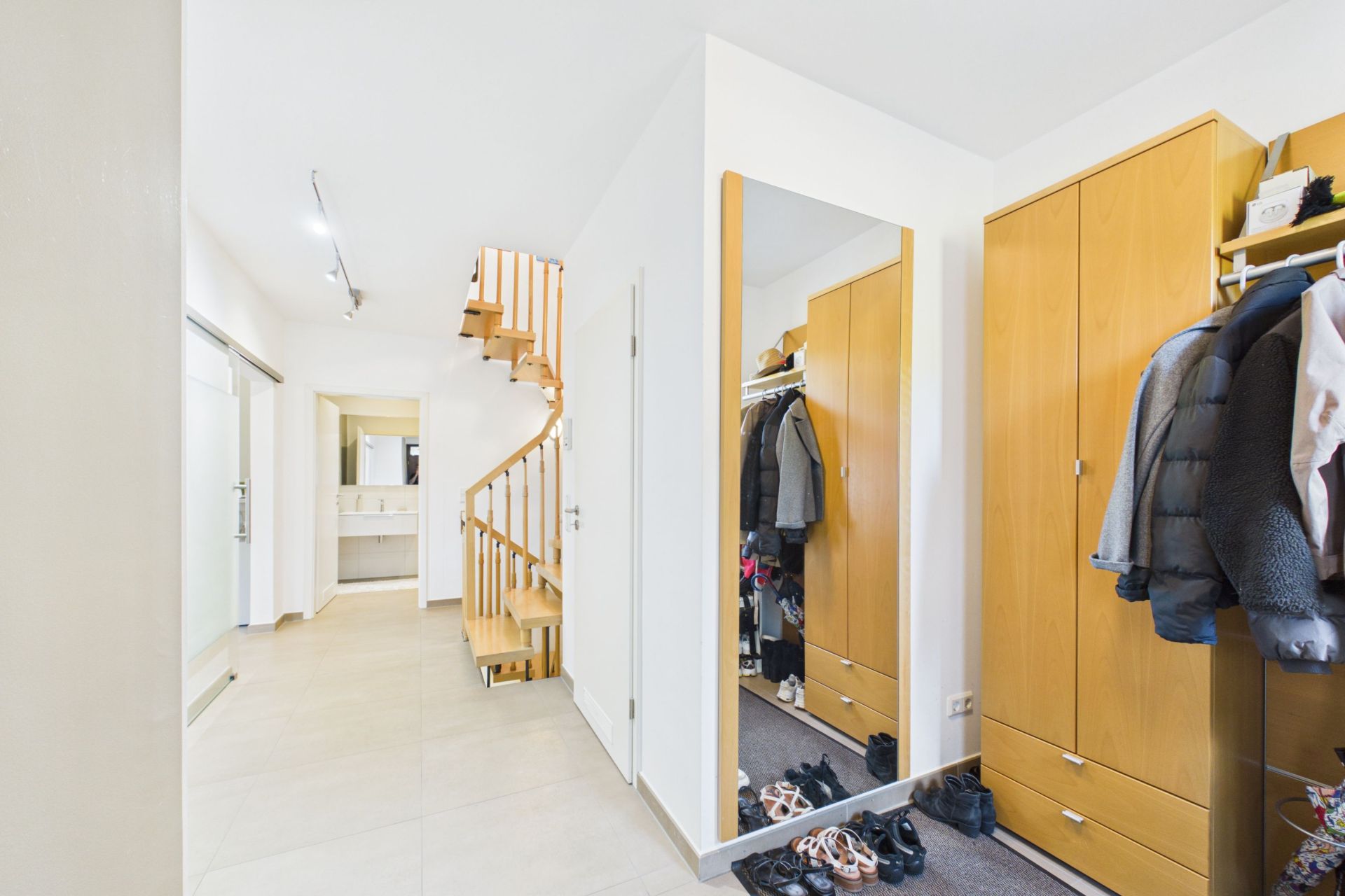 Heller Flur mit Holztreppe, Spiegel, Garderobe und Schuhen auf dem Boden.