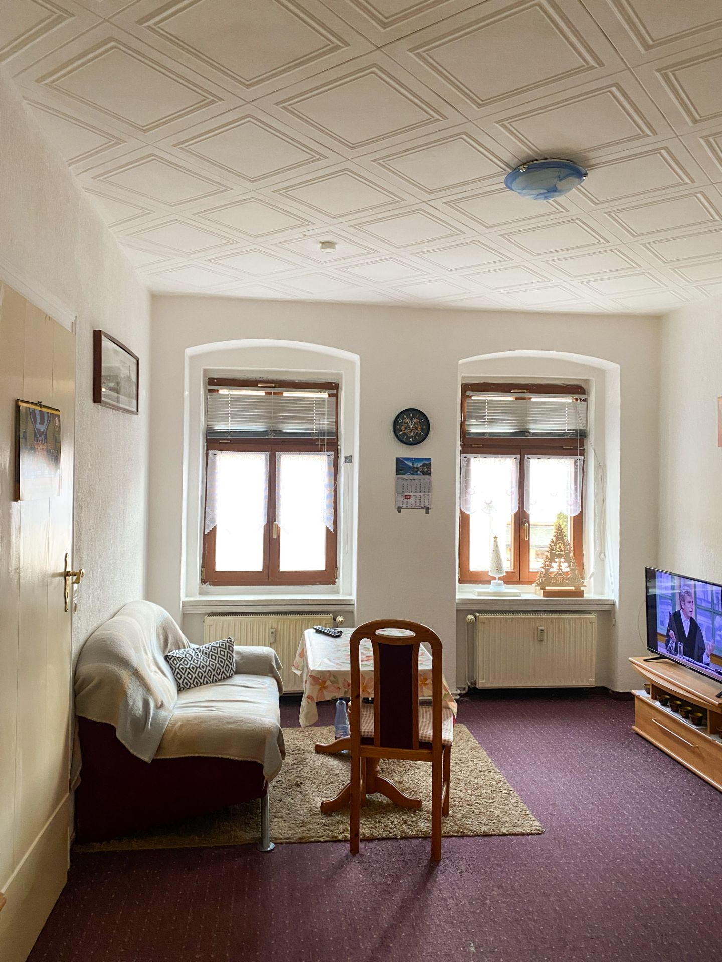 Wohnzimmer mit Sofa, Stuhl, Tisch, Teppich, Fernseher und zwei Fenstern.