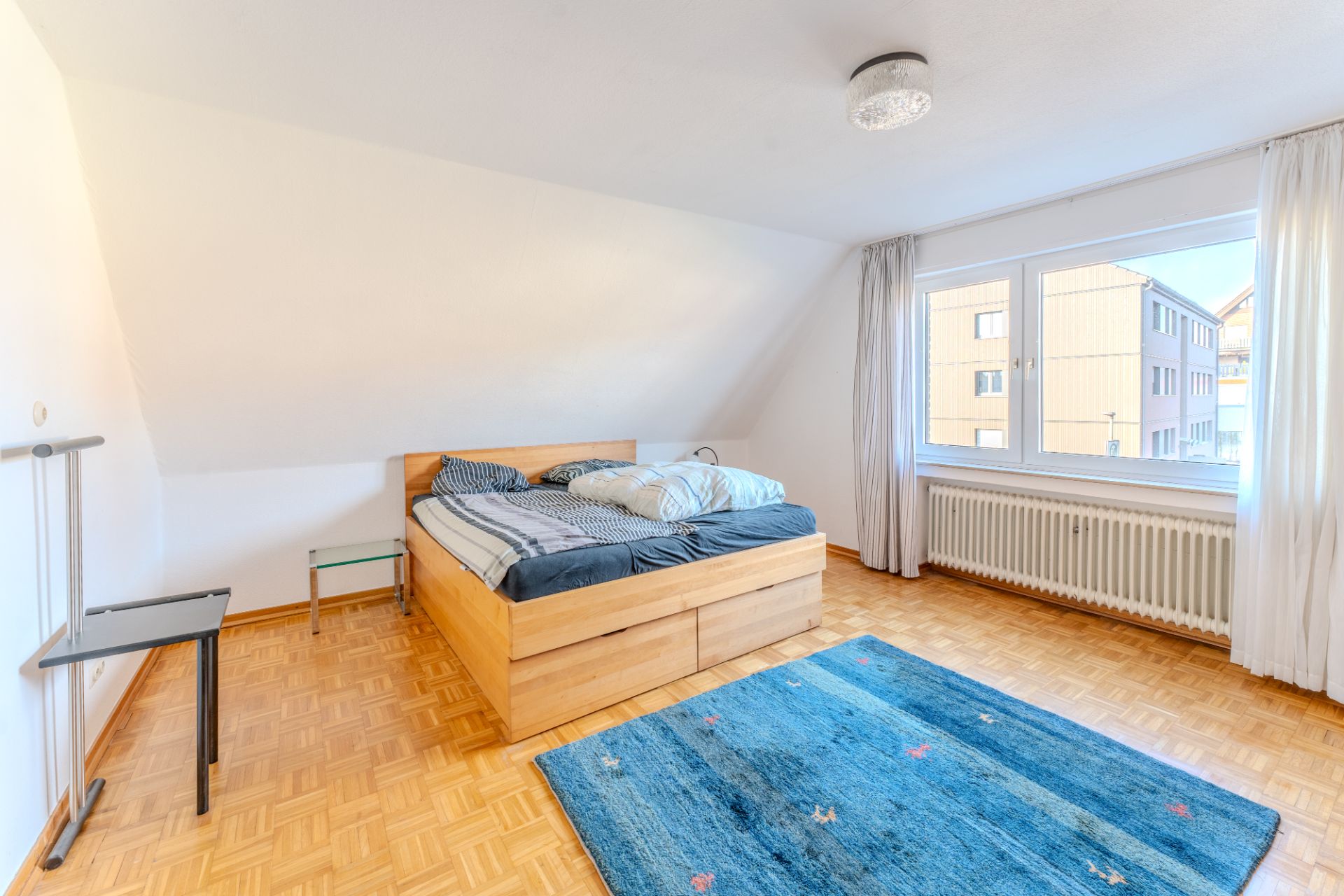 Helles Schlafzimmer mit Holzbett, blauem Teppich, großem Fenster und Parkettboden.