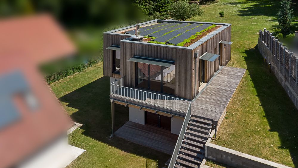 Modernes Holzhaus mit großer Terrasse, Gründach und Außentreppe auf einem grünen Grundstück.