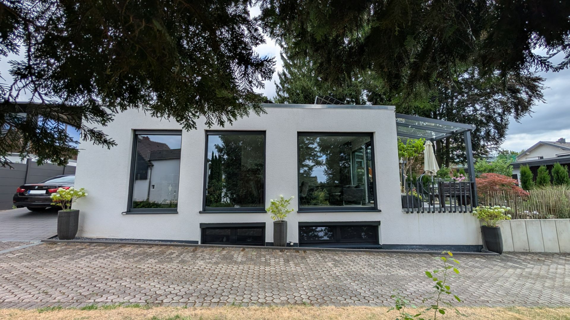 Modernes weißes Wohnhaus mit großen Fenstern, Terrasse und Pflanzkübeln, umgeben von Bäumen.