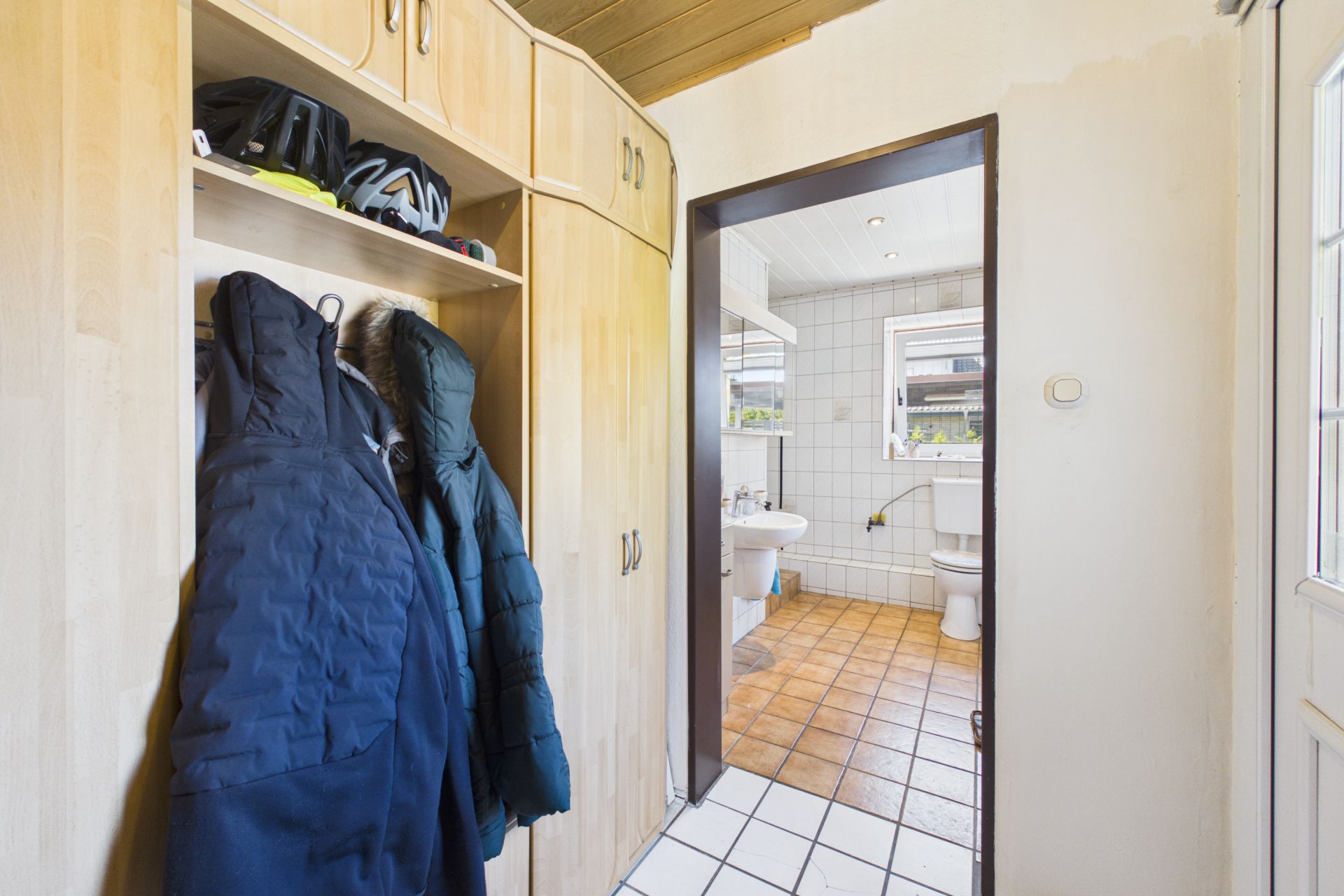 Garderobe mit Jacken und Helmen, Tür führt in ein Badezimmer mit Fliesenboden und zwei Fenstern.