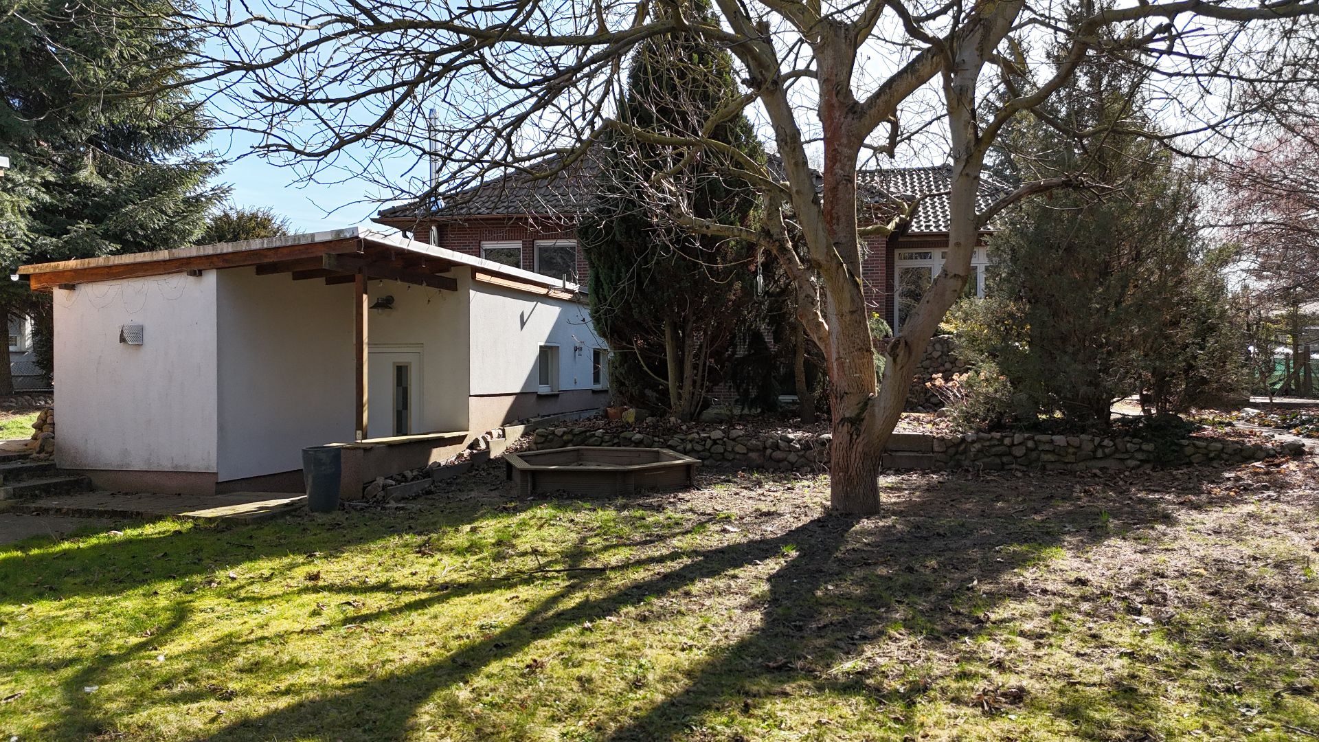 Garten mit Rasen, Baum, kleinem Nebengebäude und Wohnhaus im Hintergrund.