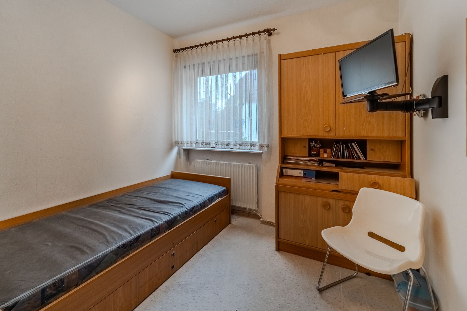 Kleines Zimmer mit Einzelbett, Holzschrank, Stuhl und Wandfernseher.