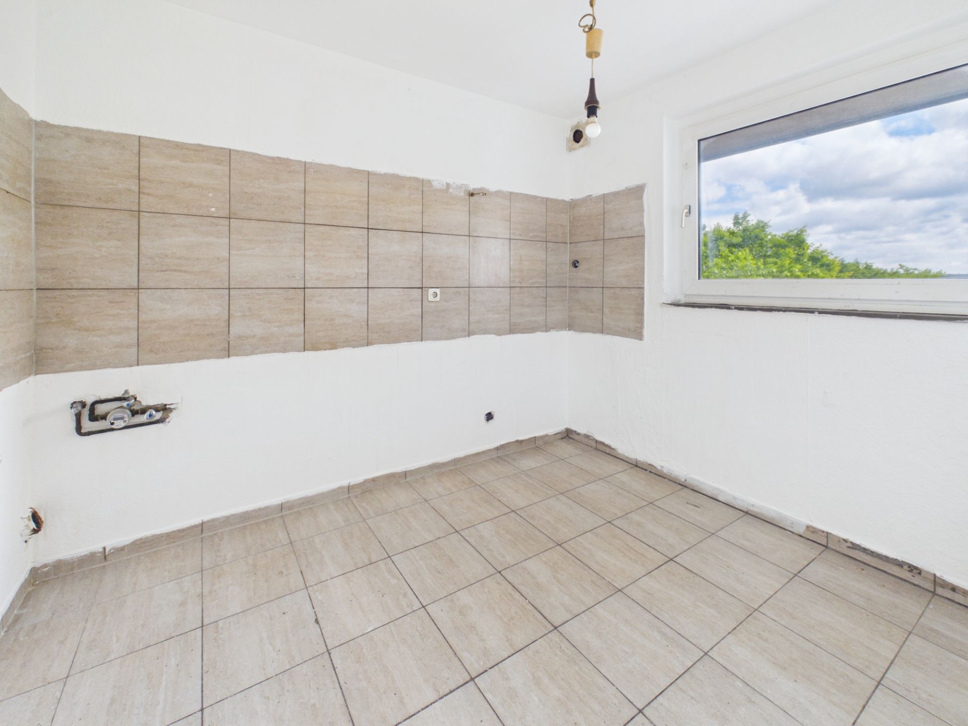 Leerer Raum mit beigen Fliesen, großem Fenster und einfacher Lampe an der Decke.