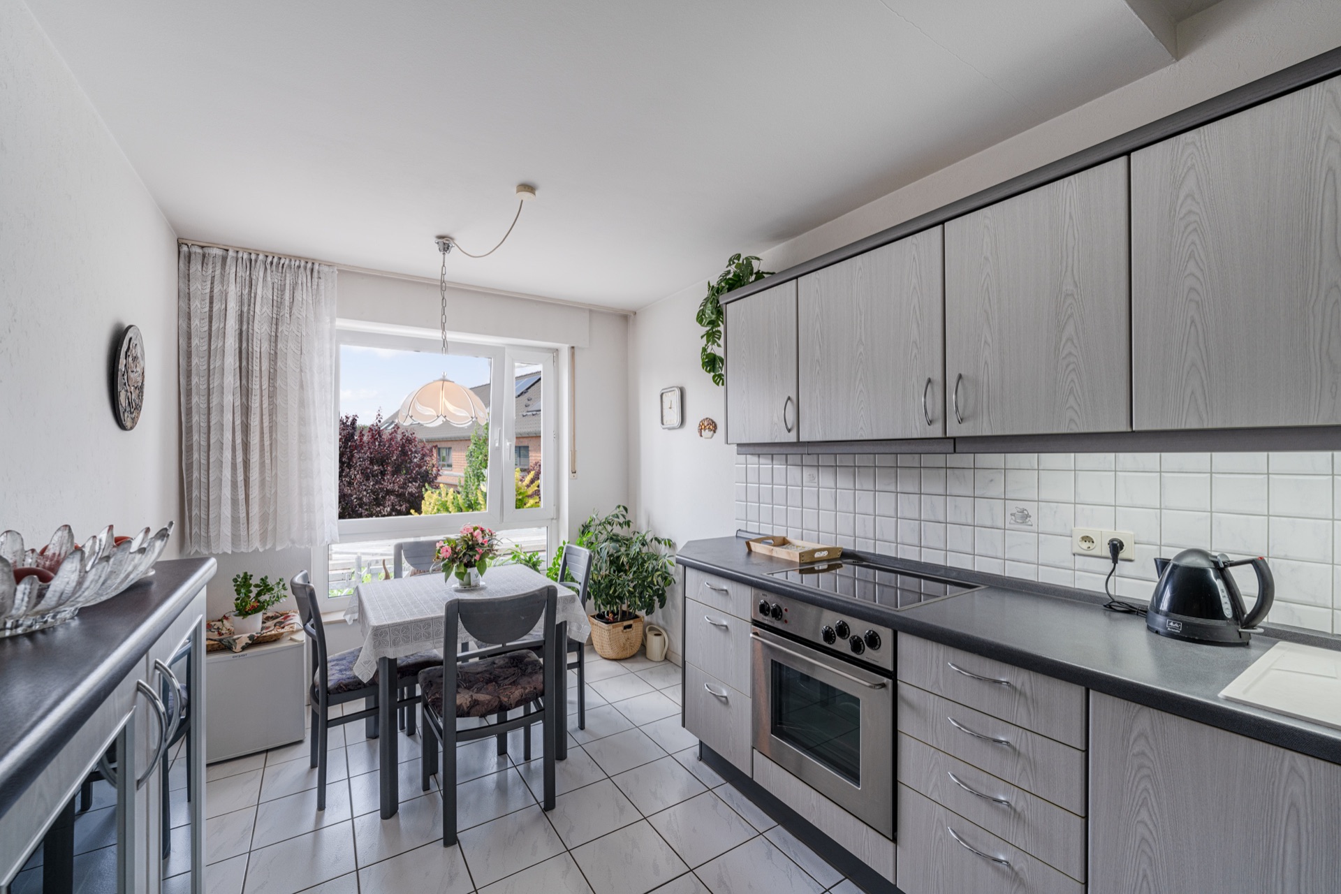 Helle Küche mit Esstisch am Fenster, Pflanzen und grauen Einbauschränken.