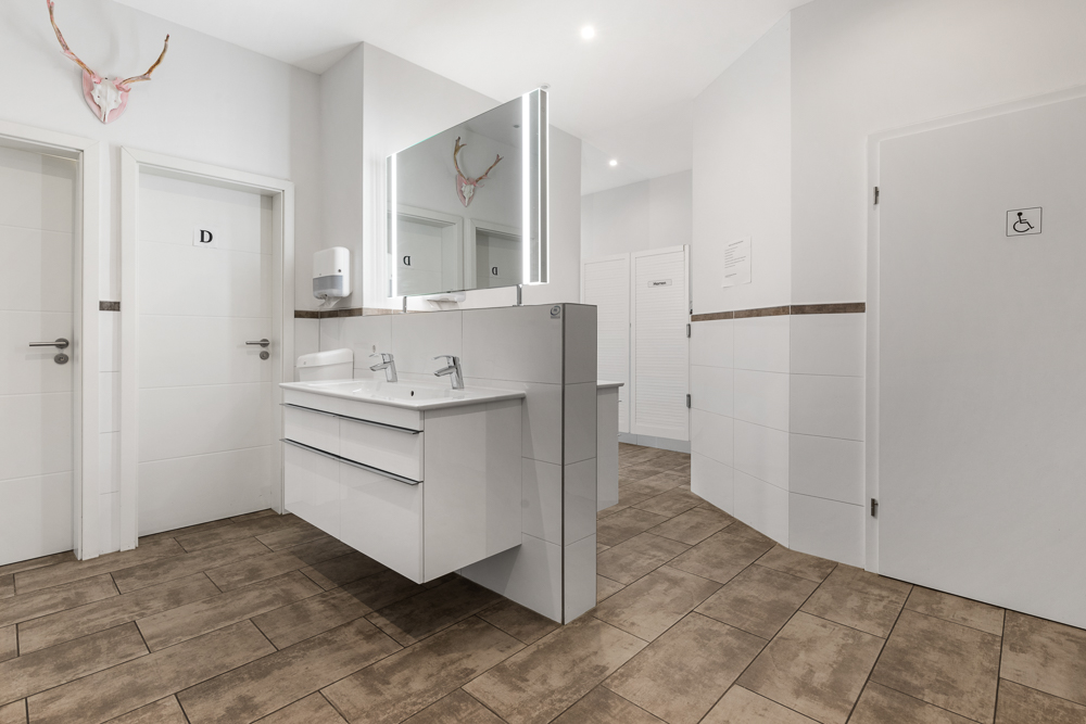 Helles Badezimmer mit zwei Waschbecken, Spiegel und mehreren Türen auf Fliesenboden.