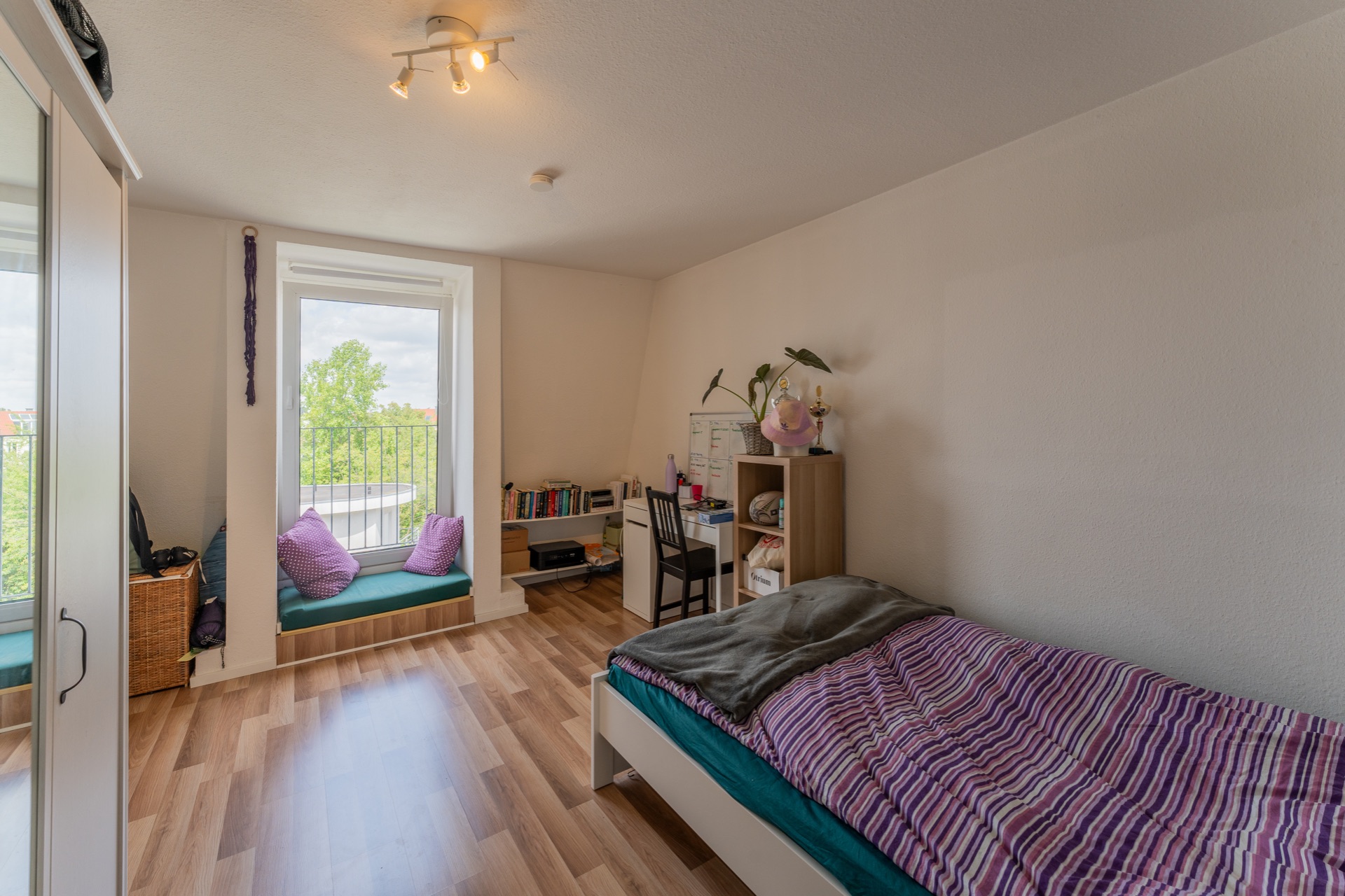 Helles Schlafzimmer mit Einzelbett, Schreibtisch, Regal und Fensterbank mit Kissen.