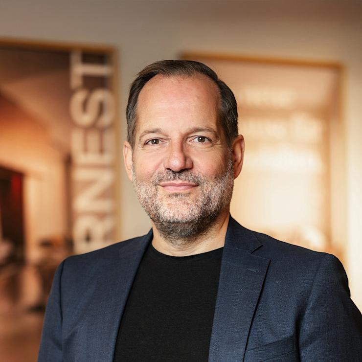 Ein Mann mit Bart trägt einen blauen Anzug und weißes Hemd, steht vor hellem Hintergrund.