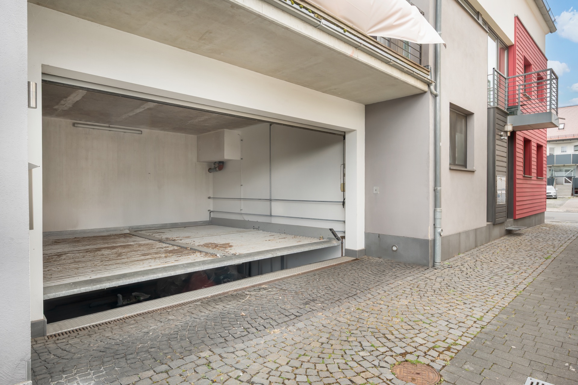 Offene Garage mit Betonboden und Decke neben einem Wohnhaus, gepflasterter Gehweg davor.