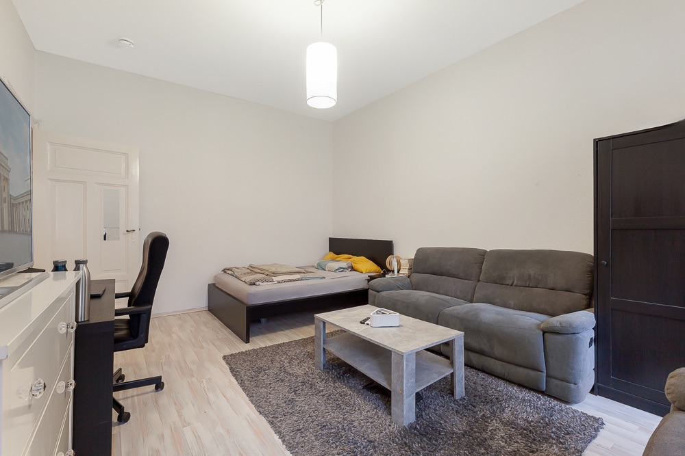 Wohnzimmer mit grauem Sofa, Bett, Schreibtisch und Teppich auf hellem Holzboden.