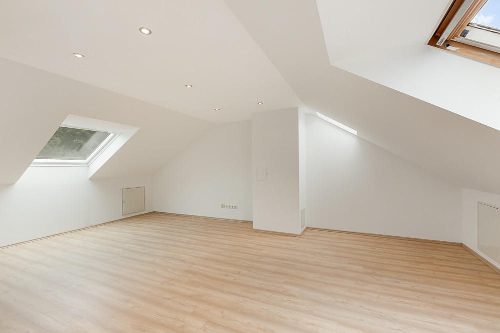 Heller Dachbodenraum mit schrägen Wänden, Holzfußboden und mehreren Dachfenstern.
