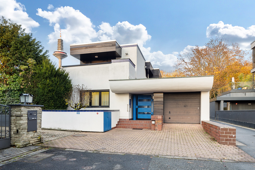 Modernes Wohnhaus mit Flachdach, Garage und gepflasterter Einfahrt, umgeben von Bäumen.