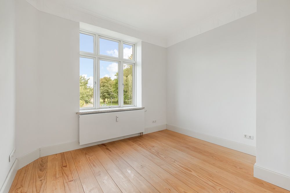 Leerer Raum mit hellem Holzboden, weißer Wand und großem Fenster mit Blick ins Grüne.