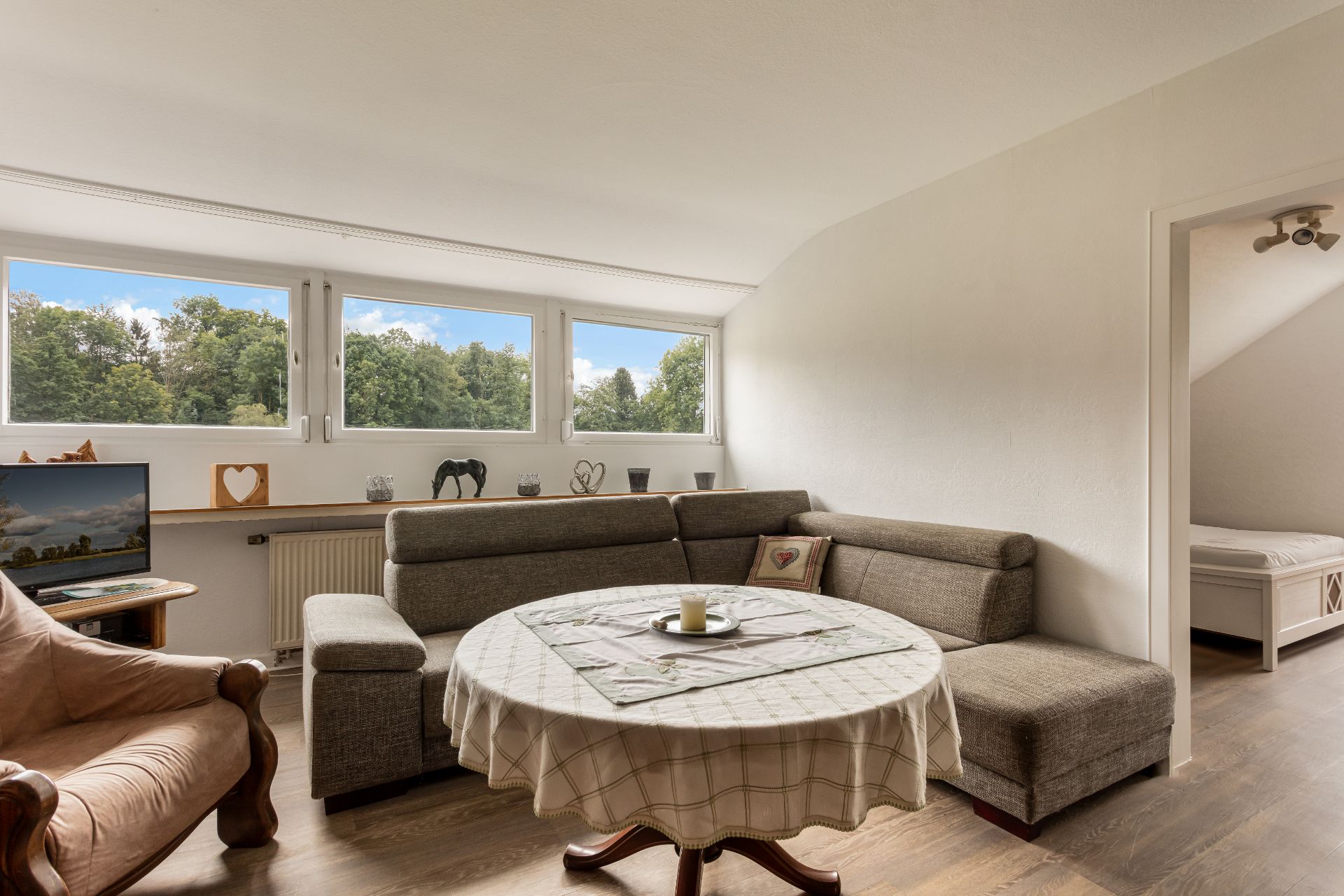 Wohnzimmer mit großer Eckcouch, rundem Tisch und Aussicht auf Bäume durch drei Fenster.