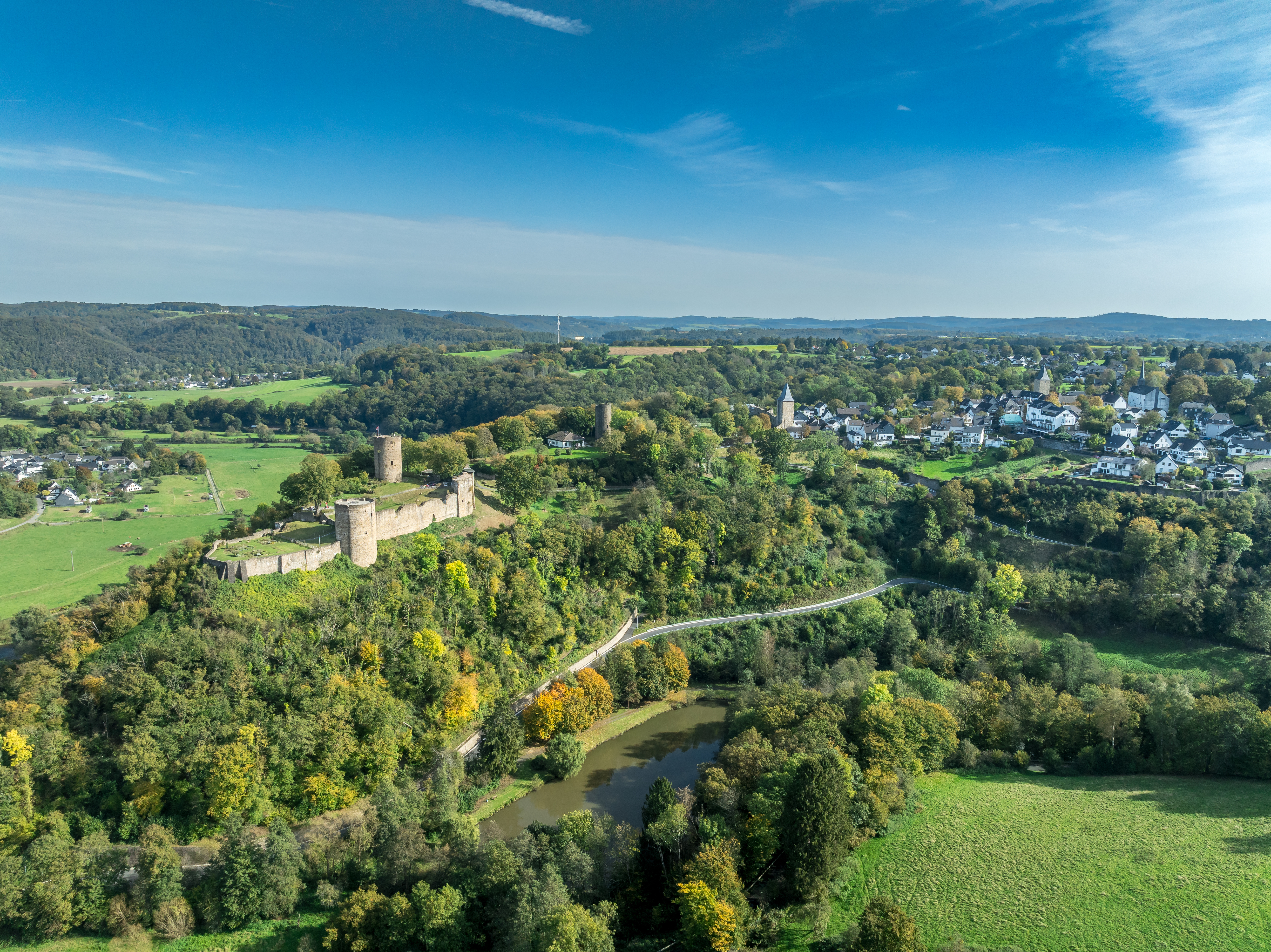 Luftaufnahme einer Burg auf einem bewaldeten Hügel mit Fluss und Dorf im Hintergrund.