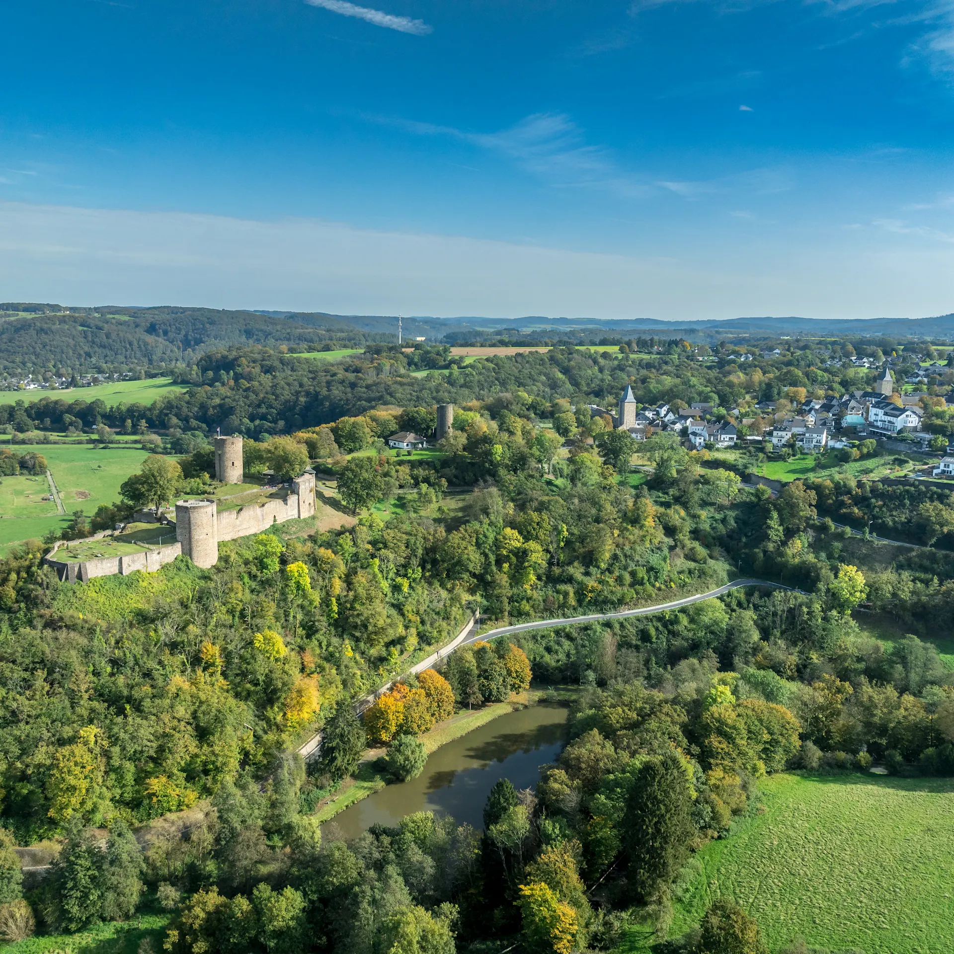 Luftaufnahme einer Burg auf einem bewaldeten Hügel mit Fluss und Dorf im Hintergrund.