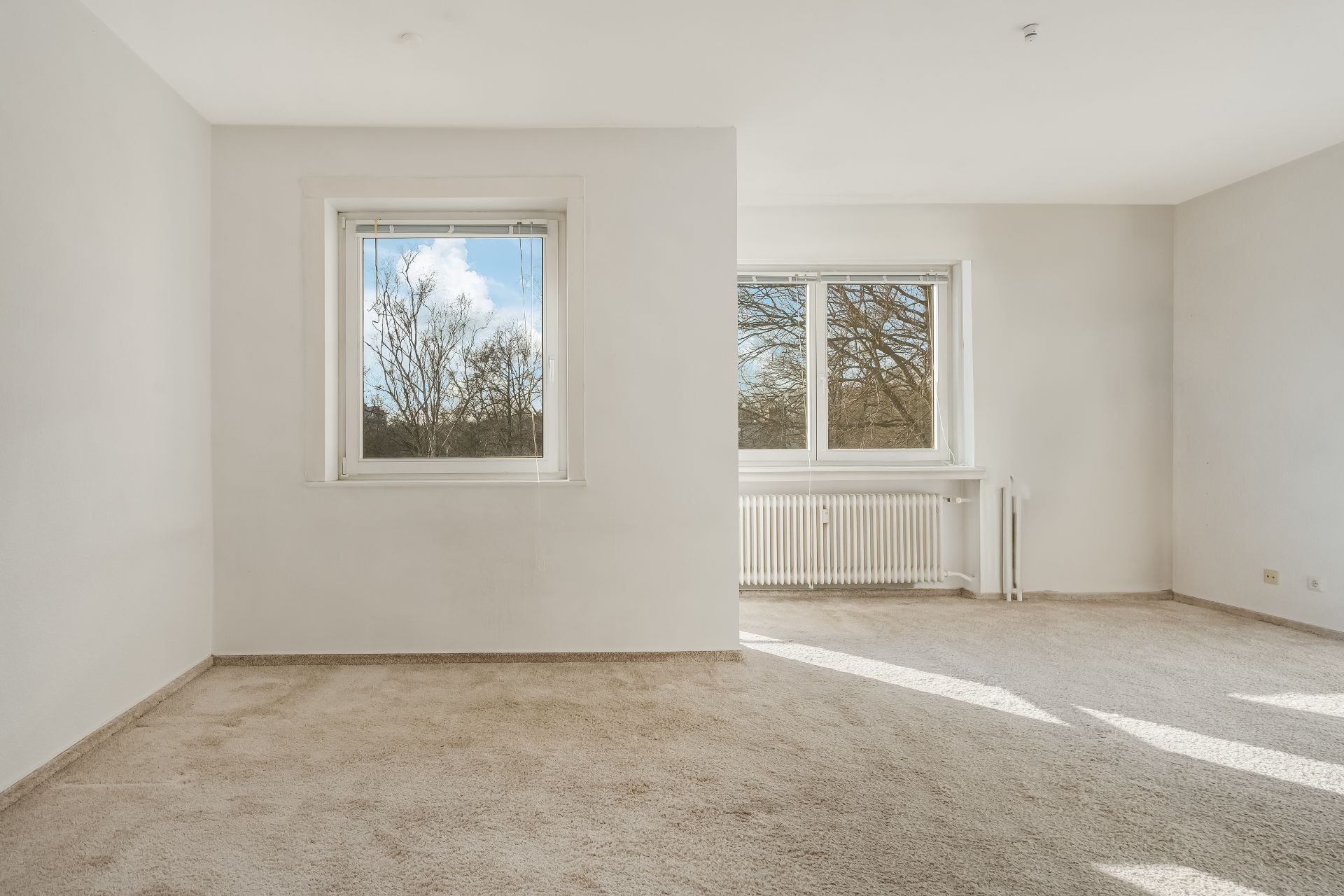 Heller, leerer Raum mit zwei Fenstern, Teppichboden und weiß gestrichenen Wänden.