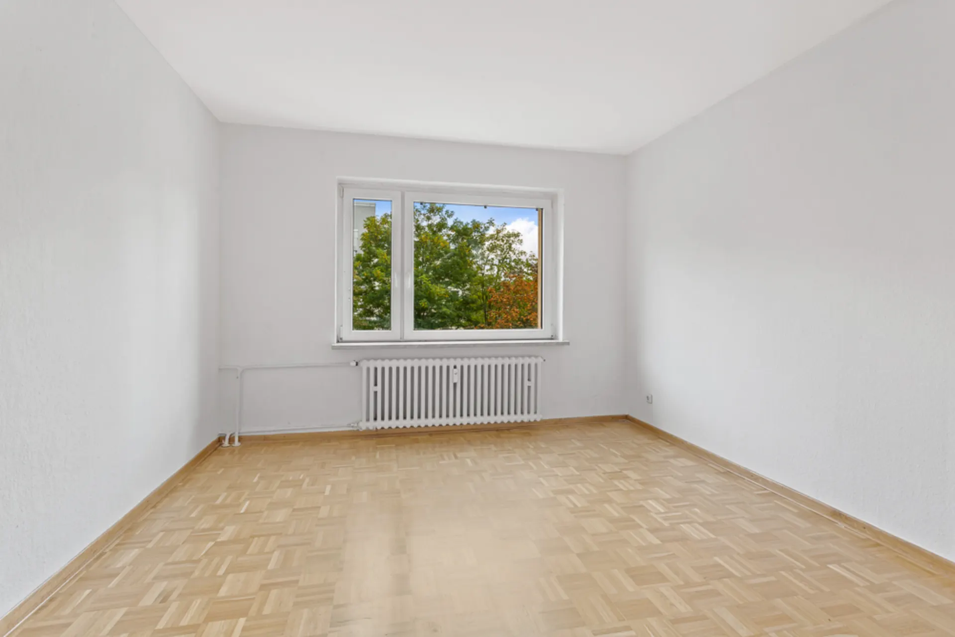Leeres Zimmer mit hellem Parkettboden, weiß gestrichenen Wänden und Fenster mit Blick ins Grüne.